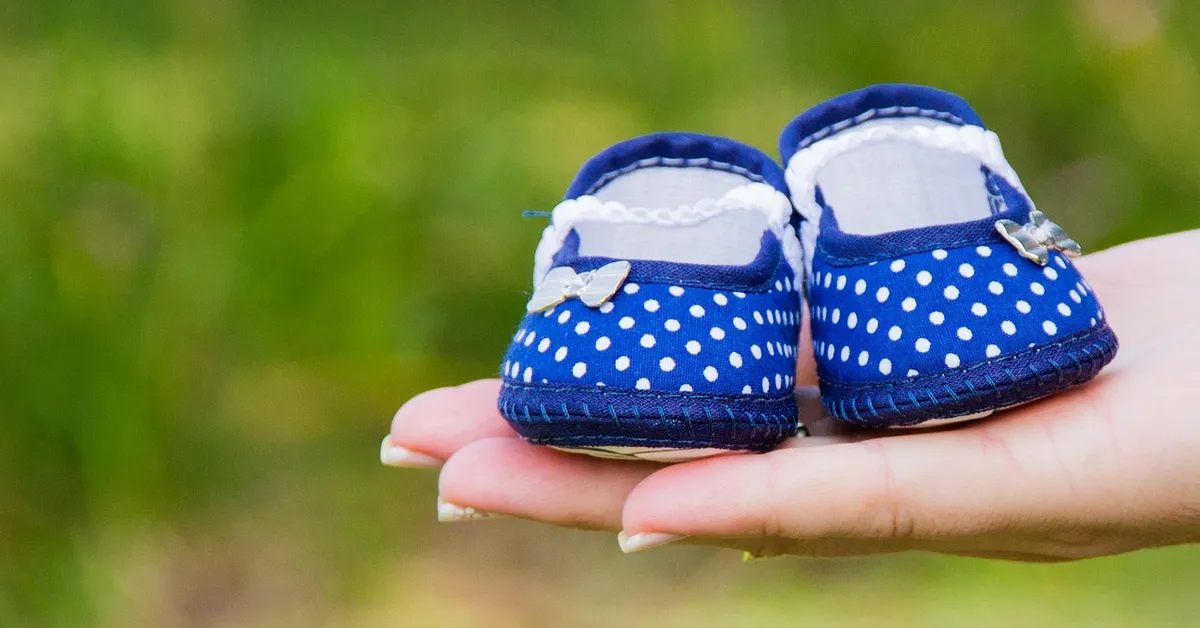 Small blue polka-dotted baby shoes resting in an adult's open hand, with a blurred green background.