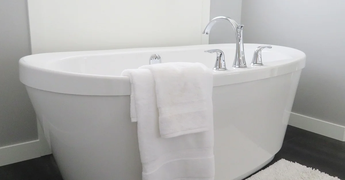 A clean white freestanding bathtub with chrome faucet in a modern bathroom, a white towel draped over the edge.