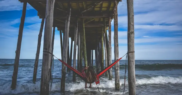Person lying in a bright red hammock tied between weathered wooden pilings under a pier at the beach.
