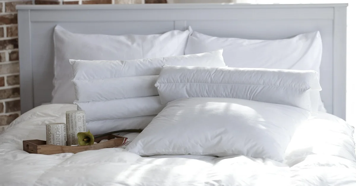 A neatly made white bed with fluffy pillows and a wooden tray on the duvet, set against a brick wall.