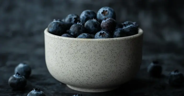 Close-up of a light speckled ceramic bowl overflowing with blueberries on a dark, moody background.