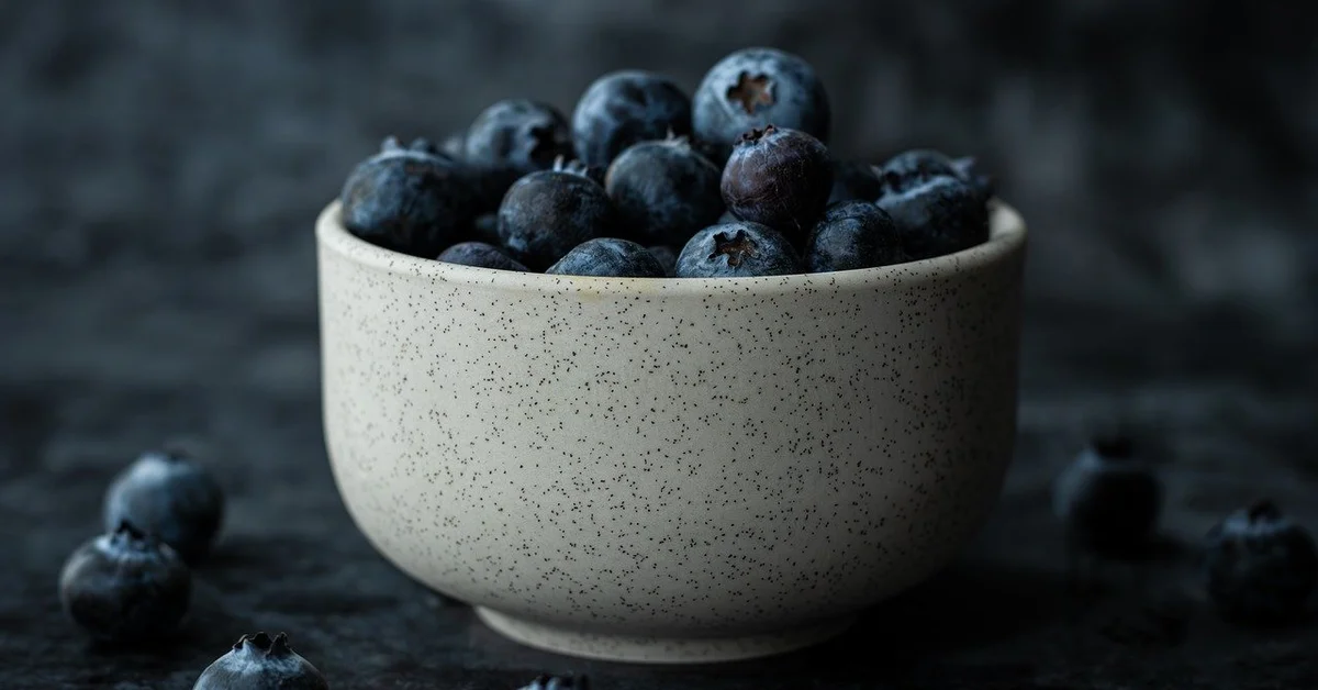 Close-up of a light speckled ceramic bowl overflowing with blueberries on a dark, moody background.
