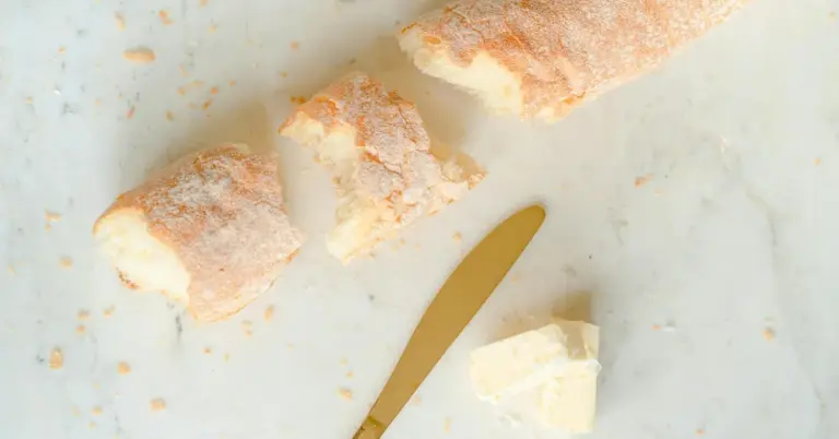 Crusty bread pieces scattered on a white marble surface with a pat of butter and a gold-colored knife.