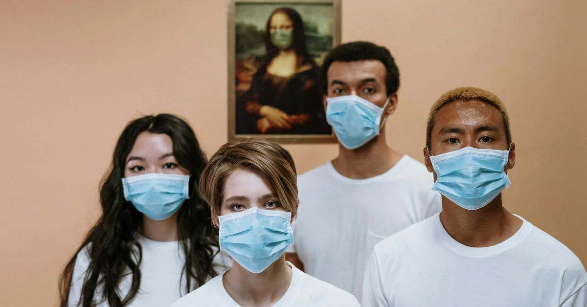 Four people wearing blue surgical masks stand in a room with a framed painting in the background.