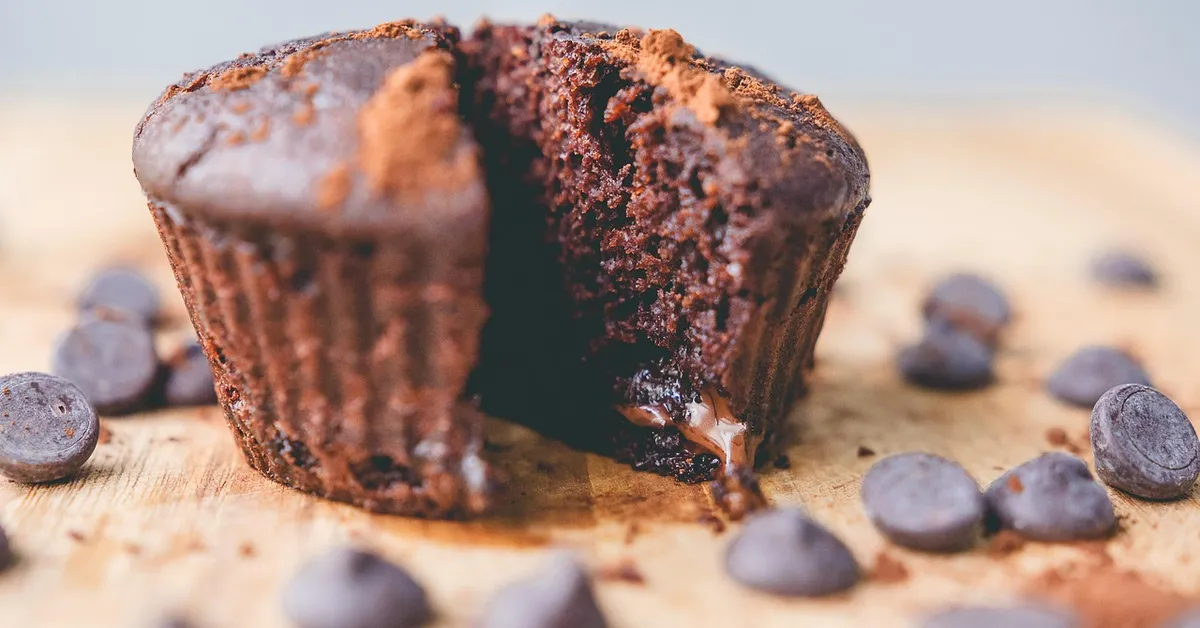 Close-up of a moist chocolate muffin with a gooey center, partially broken open, with chocolate chips scattered on a wooden surface.