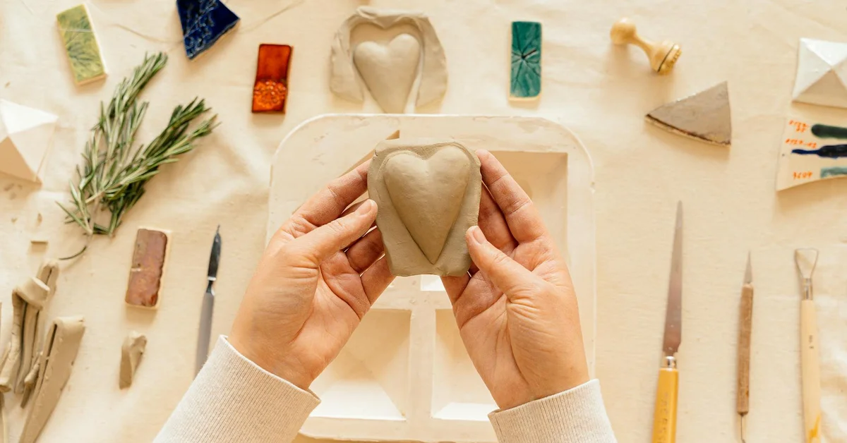 Hands shaping a clay heart on a light workspace, surrounded by pottery tools and rosemary sprigs.