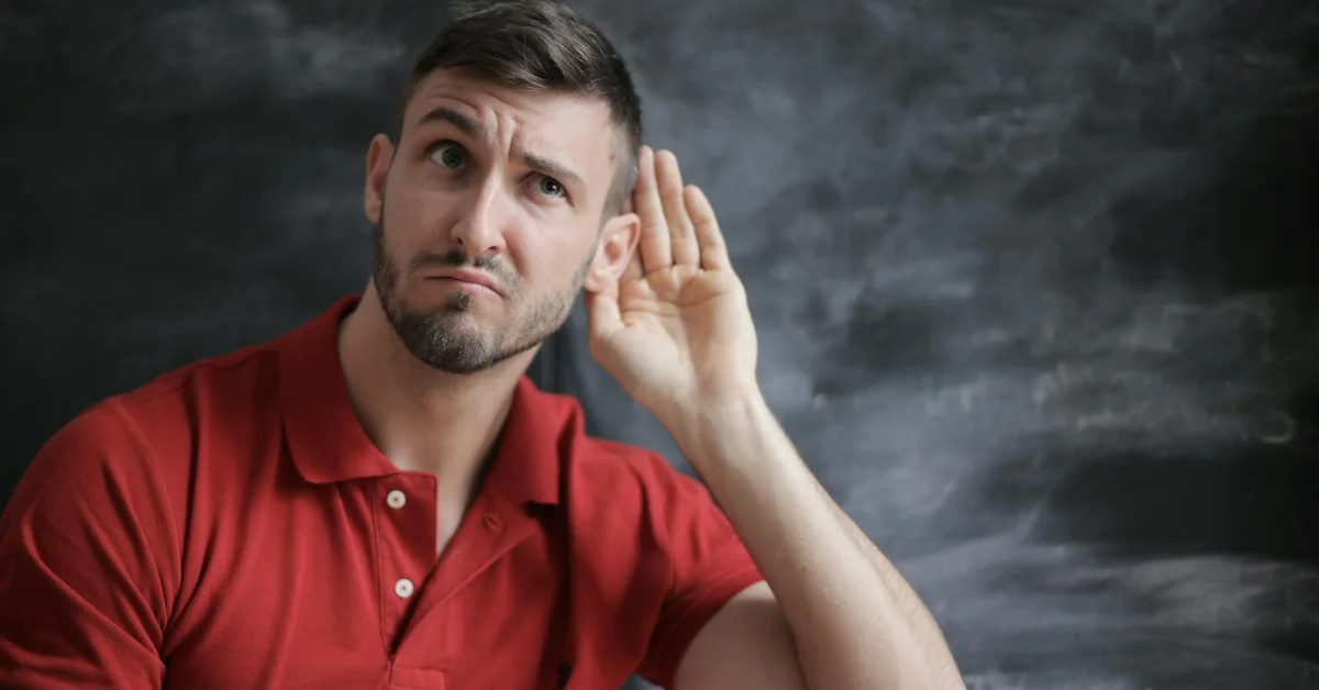 Adult man in a red polo shirt cupping his ear and tilting his head, appearing puzzled or concerned.