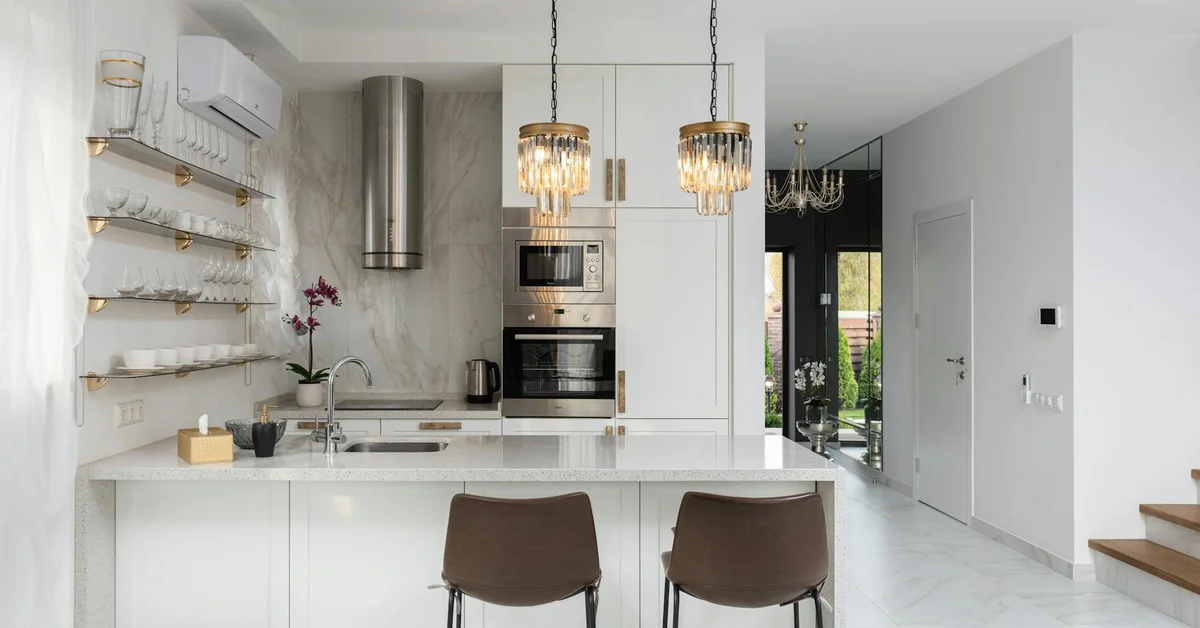 Bright modern white kitchen with an island, marble-like countertops, open shelving, and stainless steel appliances; two brown chairs sit at the island.