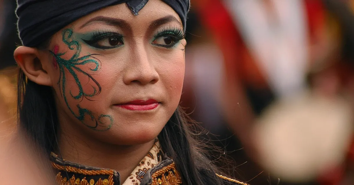 Close-up portrait of a performer with teal face paint and a decorative headband, looking calmly to the side.