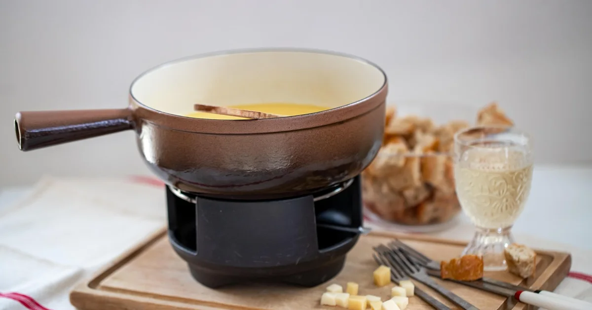 A brown fondue pot holding melted cheese sits atop a stand, with cubed cheese and pieces of bread on a wooden board nearby, illustrating foods to consider as bait for a pet mouse.