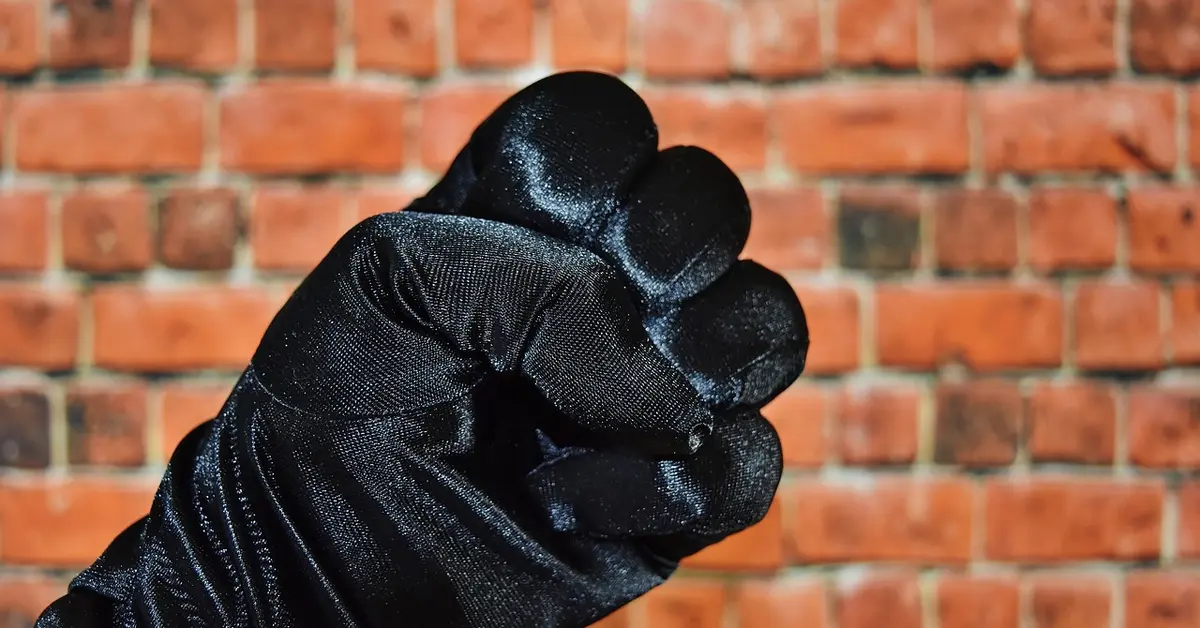 A gloved hand forming a fist in a threatening gesture against a brick wall.