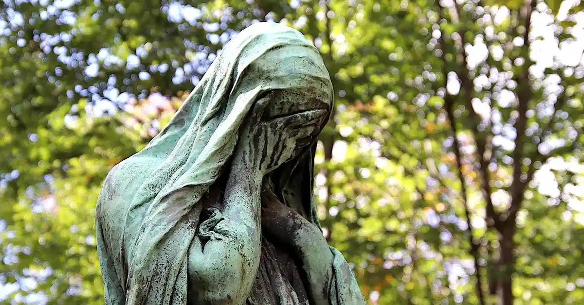 Bronze-green statue of a grieving figure in a sunlit park, with leafy trees in the background.