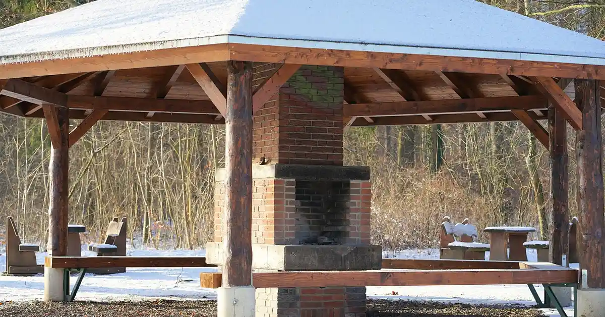 Outdoor brick barbecue grill under a wooden pavilion with snow on the roof, surrounded by picnic tables