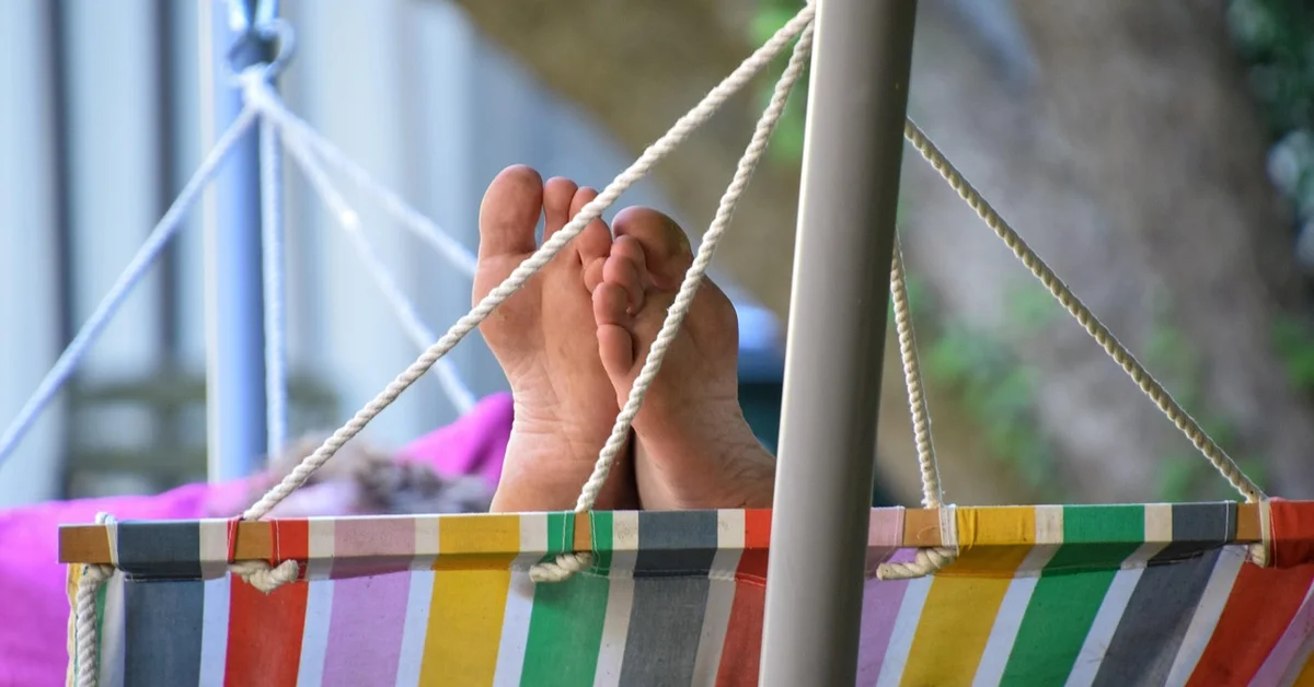 A person relaxing in a colorful striped hammock outdoors, with bare feet propped up and ropes securing the hammock to a frame.