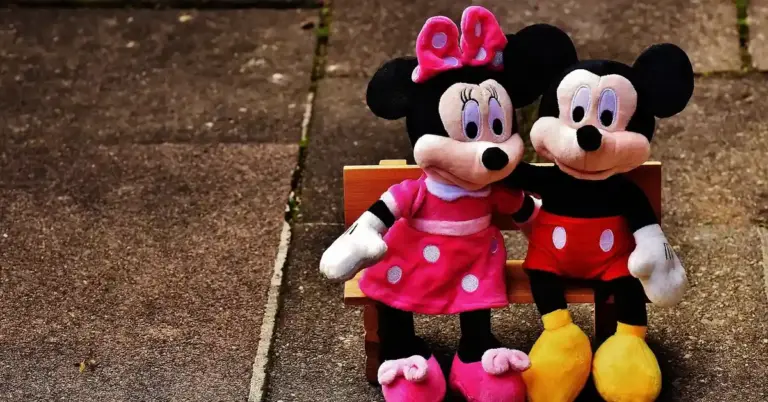Plush Mickey and Minnie Mouse dolls sitting together on a wooden bench outdoors