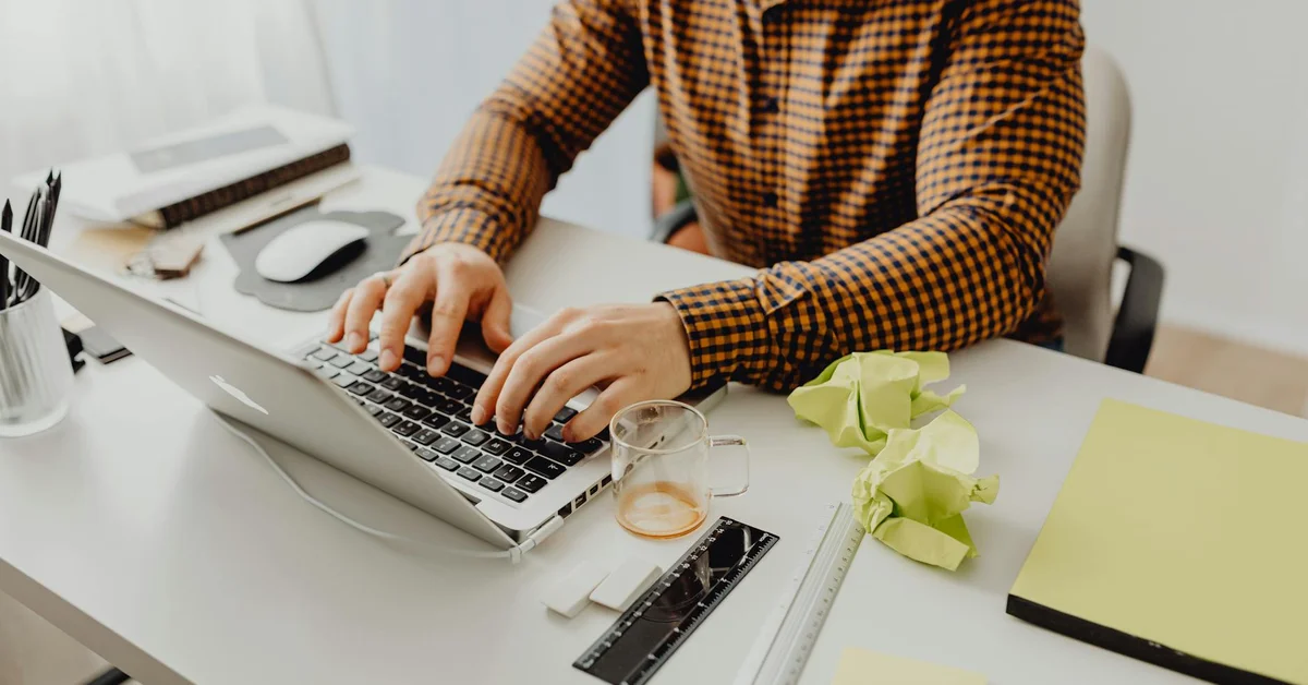 A person working on a laptop at a desk with a unique color palette, featuring a checkered shirt and bright yellow materials.