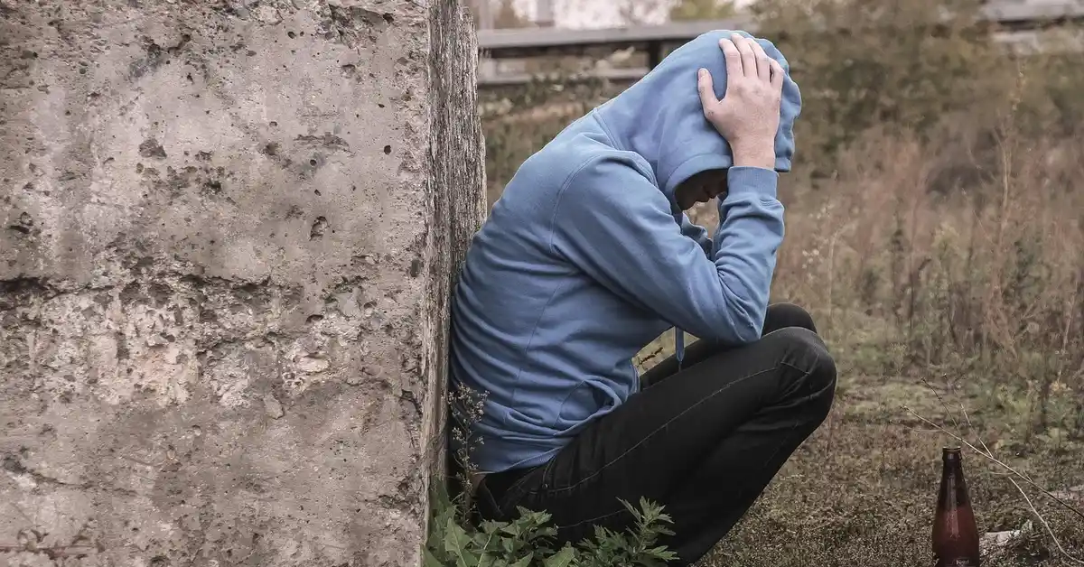 A person in a blue hoodie sits against a rough concrete wall outdoors, head in hands, appearing distressed and exhausted.