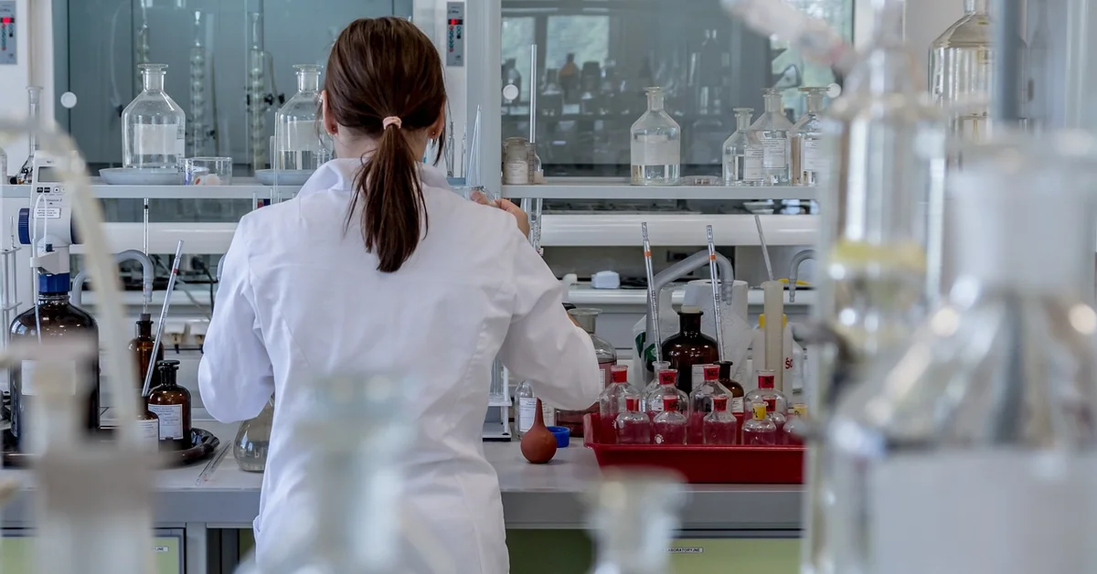 A laboratory technician in a white coat works at a bench filled with glassware and chemical bottles, representing the controlled environment used for researching mice.