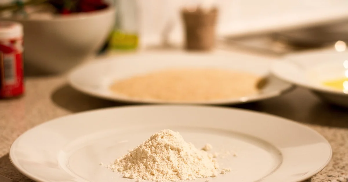 A mound of flour on a white plate on a kitchen counter with blurred bowls and kitchen items in the background.
