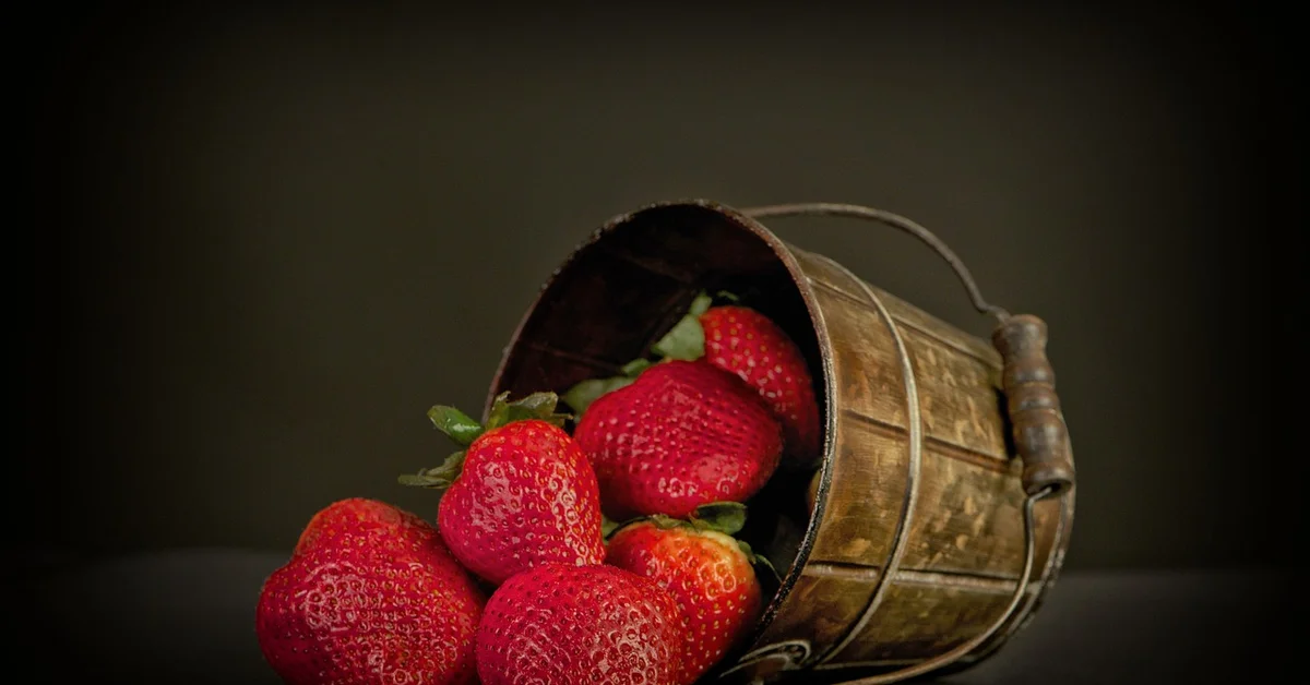A rustic wooden bucket tipped on its side with bright red strawberries spilling onto a dark surface.