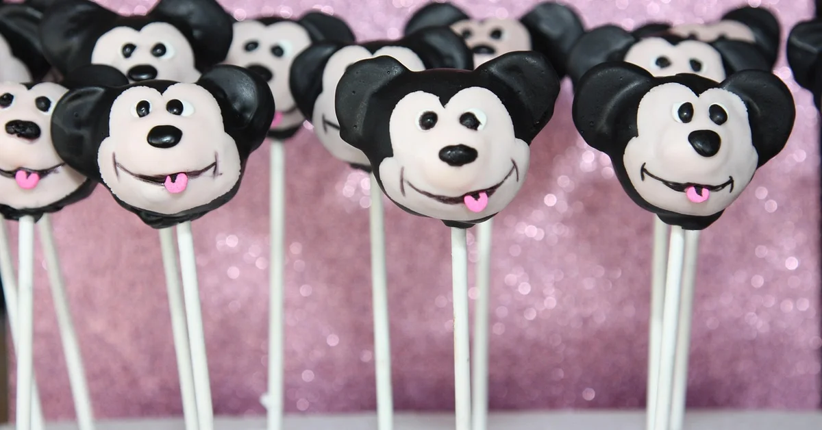 A row of Mickey Mouse head-shaped lollipops on sticks against a pink glittery background.