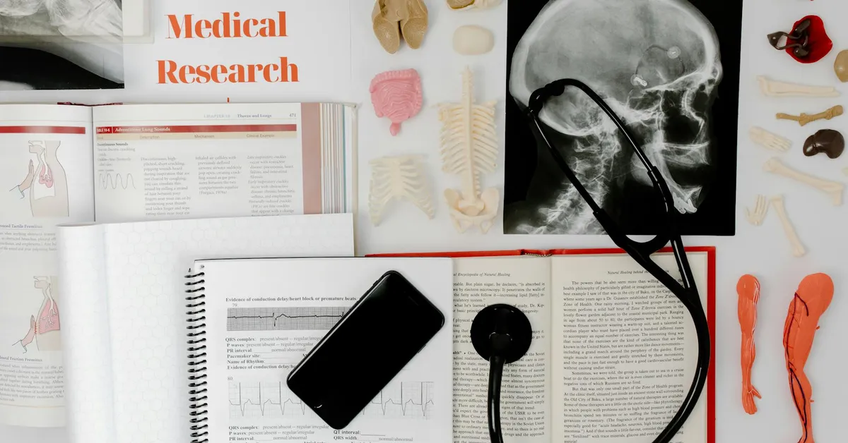 Flat lay of medical anatomy resources: an X‑ray of a skull with a stethoscope, a medical research sign, open textbooks and notebooks with charts, a smartphone, and plastic bone models, illustrating reference materials for studying mouse skeletal anatomy.