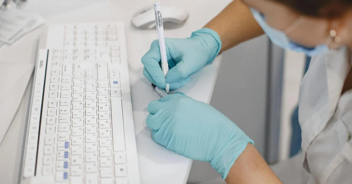 A researcher wearing gloves looks closely at a mouse sample while taking notes in a laboratory setting.