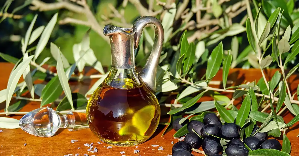 A glass olive oil bottle with a curved handle and spout on a wooden surface, surrounded by olive leaves and black olives