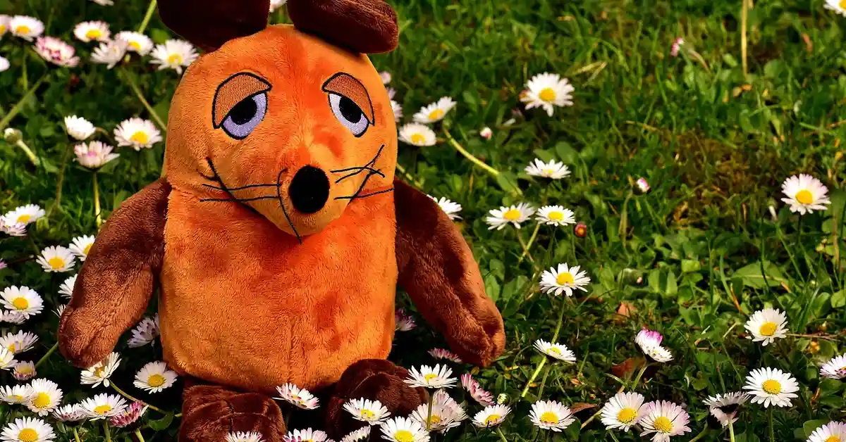 A soft orange plush mouse with brown ears and limbs sits among white daisies in a sunlit field.