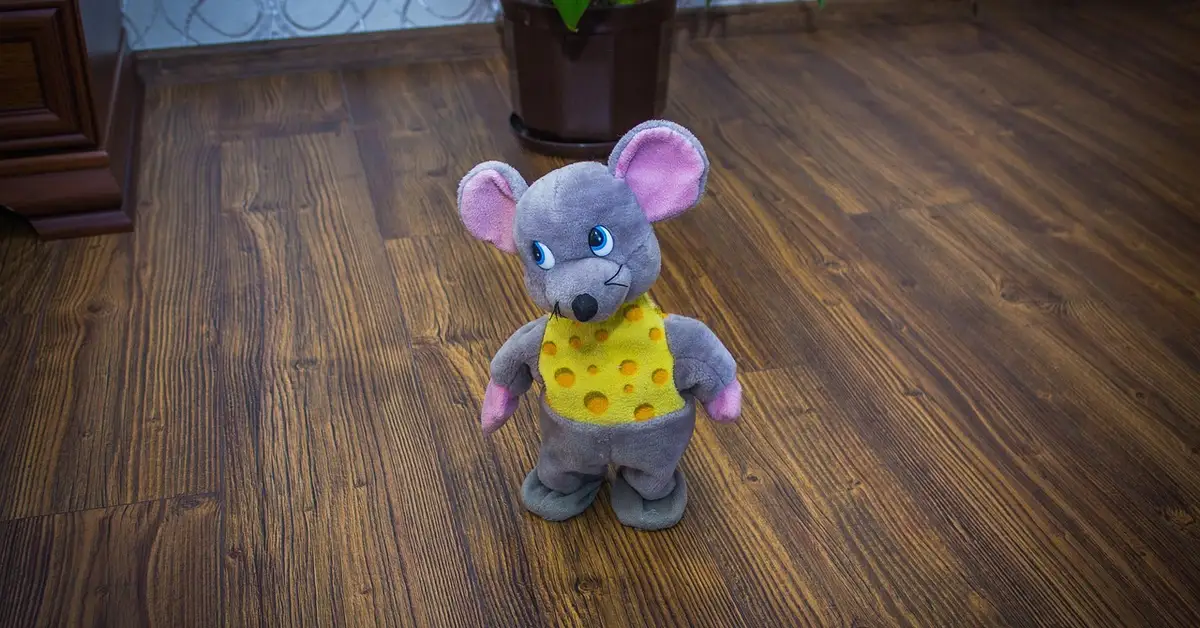 Soft gray plush mouse with pink ears wearing a yellow cheese-patterned vest, standing on a wooden floor.