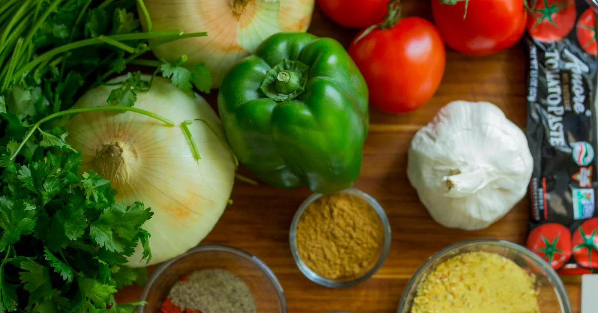 Fresh vegetables including onions, a green bell pepper, tomatoes, garlic, cilantro, parsley, and spices arranged on a wooden surface.