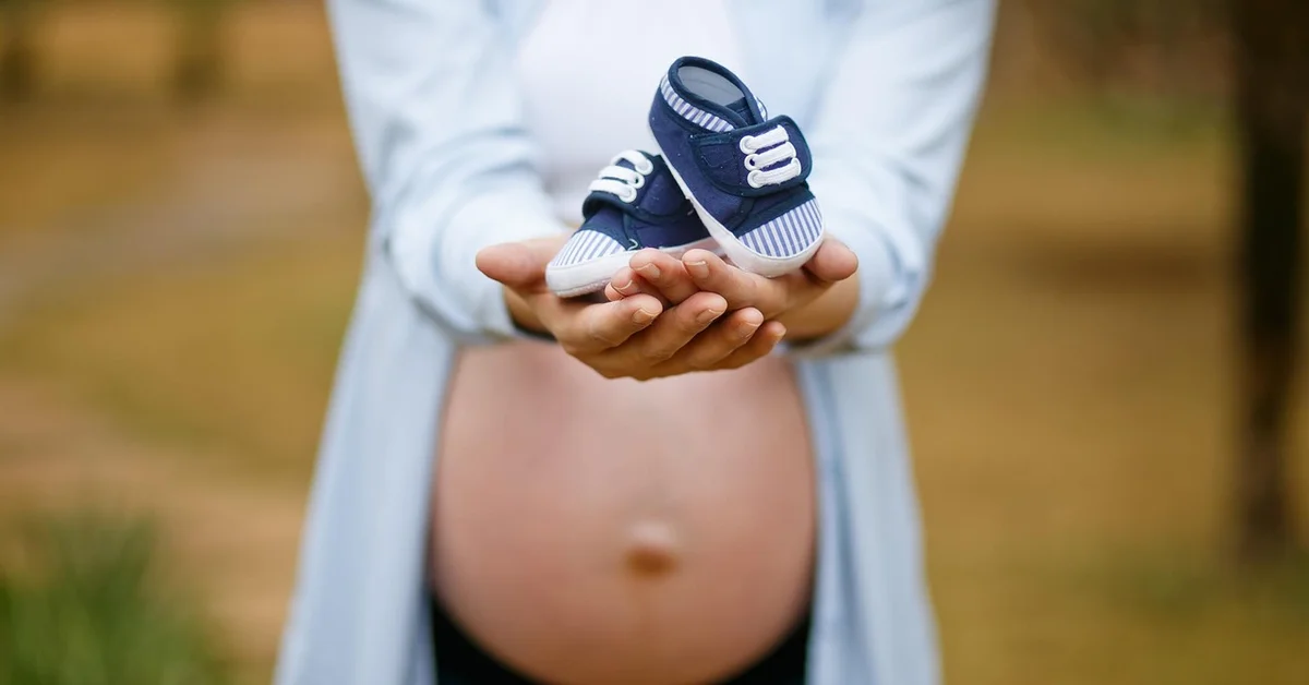 A pregnant person holding a small pair of blue baby shoes in their hands, with a blurred outdoor background.