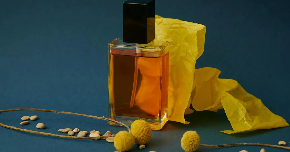 A still-life composition featuring a glass perfume bottle with amber liquid, yellow tissue paper, dried stems, and small beige seeds set against a deep blue background, evoking olfactory cues involved in scent marking.