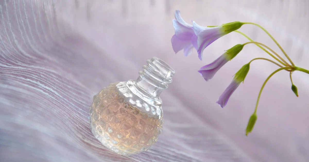 A textured glass perfume bottle resting on soft pink fabric beside a cluster of delicate purple flowers, illustrating scent and aroma cues.