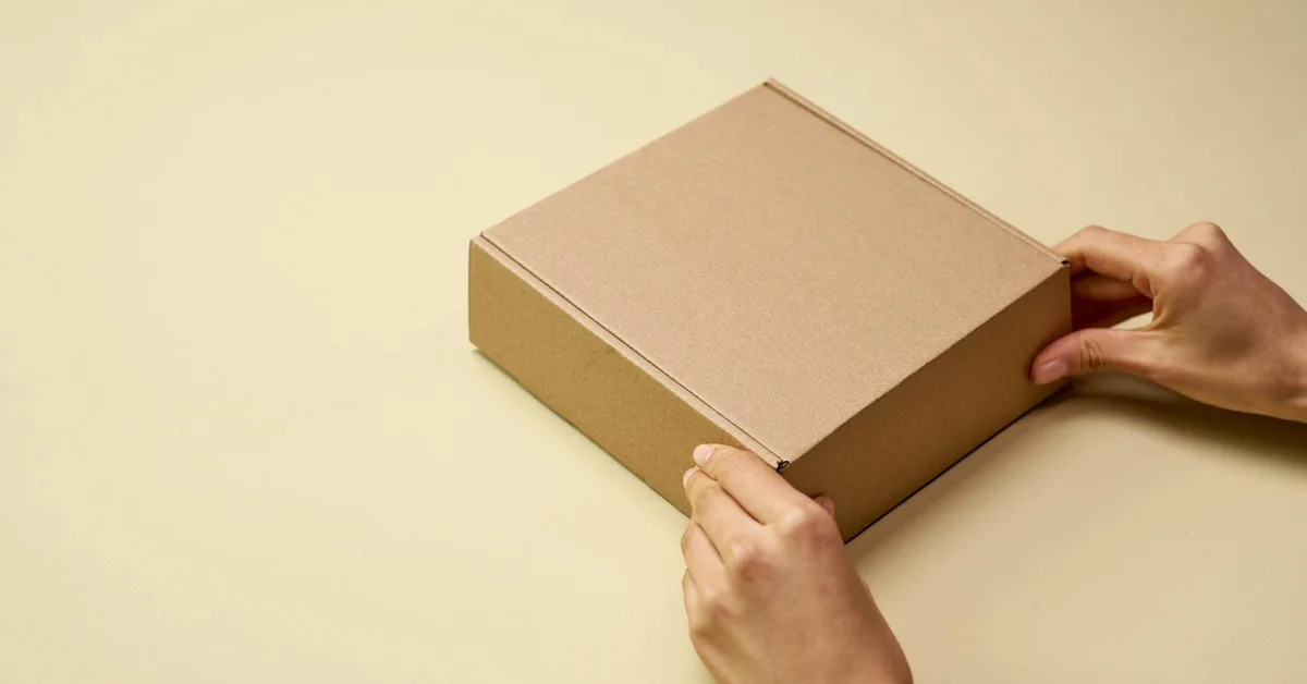 Person's hands assembling a plain cardboard box on a light, neutral surface.