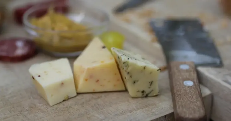 Three wedges of cheese on a wooden cutting board with a knife in the foreground and a blurred background of kitchen items.