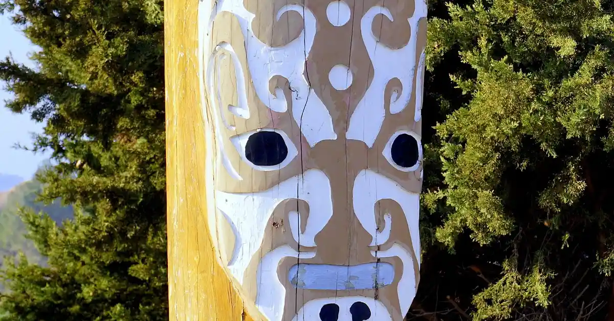 Carved wooden totem mask with brown and white patterns mounted on a vertical wooden post, surrounded by green bushes.