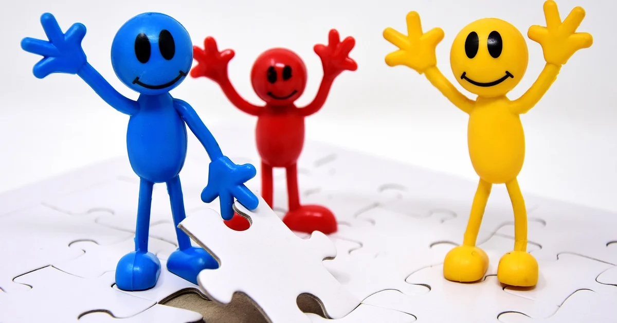 Three colorful cartoon-style toy figures (blue, red, and yellow) stand on a white puzzle surface, smiling and raising their hands as puzzle pieces lie around.