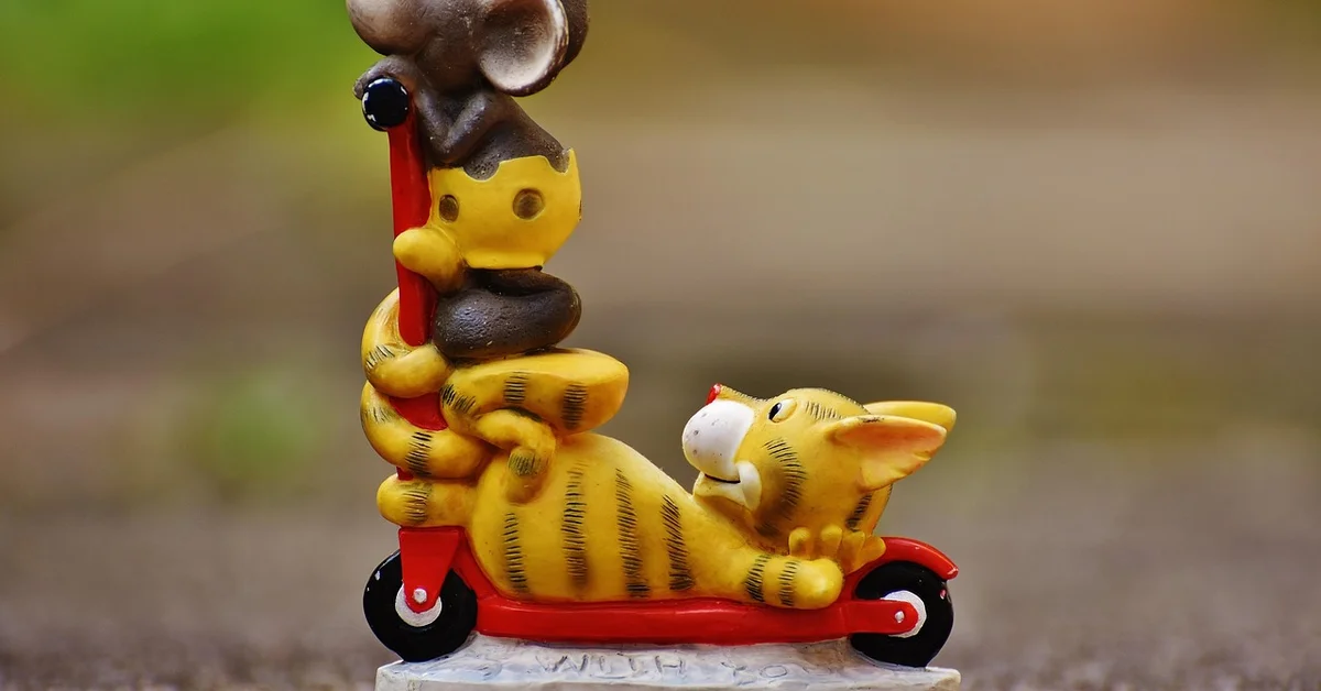 A toy scene featuring a yellow striped tiger lounging on a red toy scooter, with a small mouse figure standing on the scooter's handlebar.
