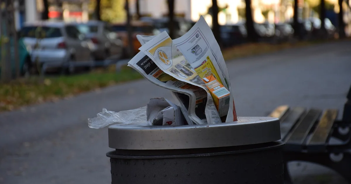 A metal outdoor trash can with crumpled newspapers sticking out, placed on a park-like walkway with blurred trees and parked cars in the background.