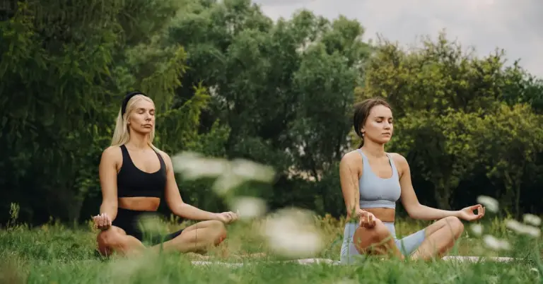 Two women sit cross-legged on yoga mats in a green park, meditating with closed eyes and relaxed posture.