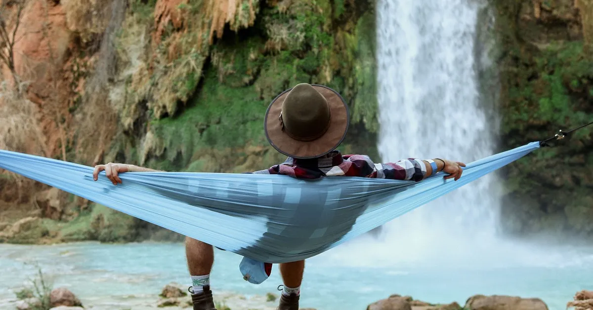 A person wearing a wide-brimmed hat lounges in a light blue hammock stretched between two anchor points by a waterfall.