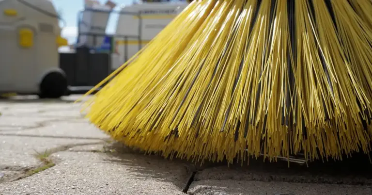 Close-up of a yellow outdoor broom resting on a paved surface with blurred cleaning equipment in the background.