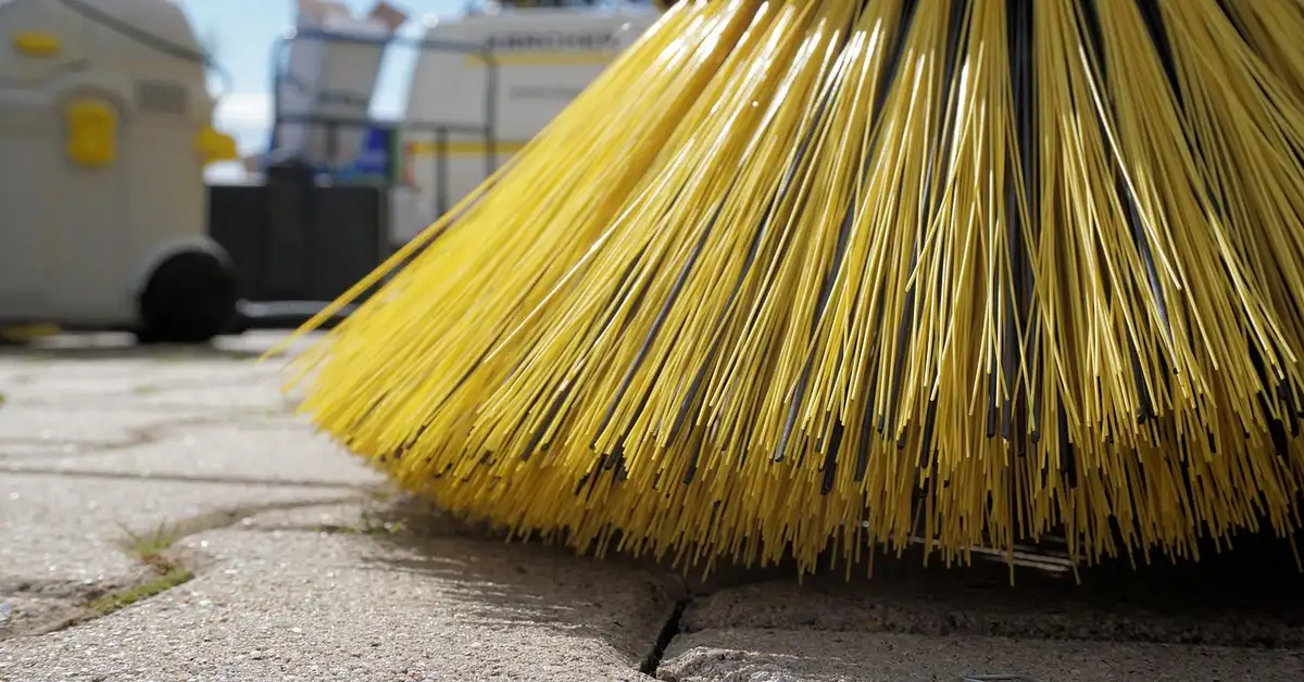 Close-up of a yellow outdoor broom resting on a paved surface with blurred cleaning equipment in the background.